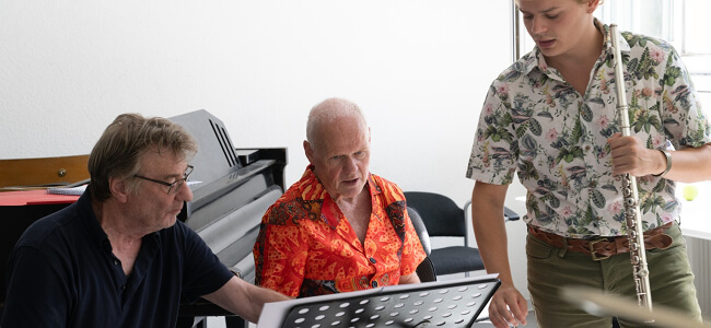 Ein Foto mit zwei Musikern vom Ensemble Modern und einem jungen Flöten-Schüler.
Die Musiker arbeiten zusammen mit dem Schüler an einem Musik-Stück.