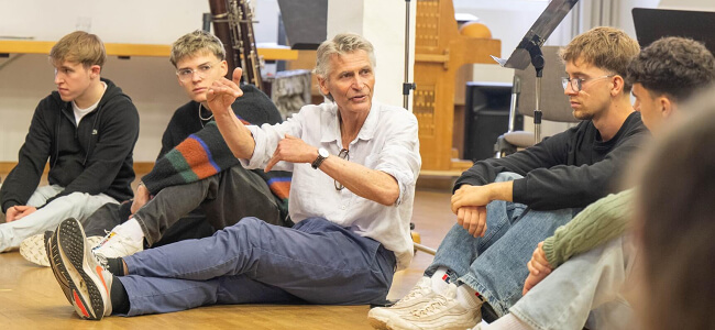 Ein Foto mit einem Musiker vom Ensemble Modern und mehreren Schülern.
Alle Personen sitzen auf dem Boden.
Der Musiker erklärt den Schülern etwas.