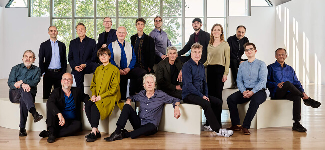 Ein Gruppen-Foto mit 17 Musiker*innen vom Ensemble Modern.
Die Musiker*innen stehen oder sitzen in einem Saal.
Sie sind sehr schick angezogen.